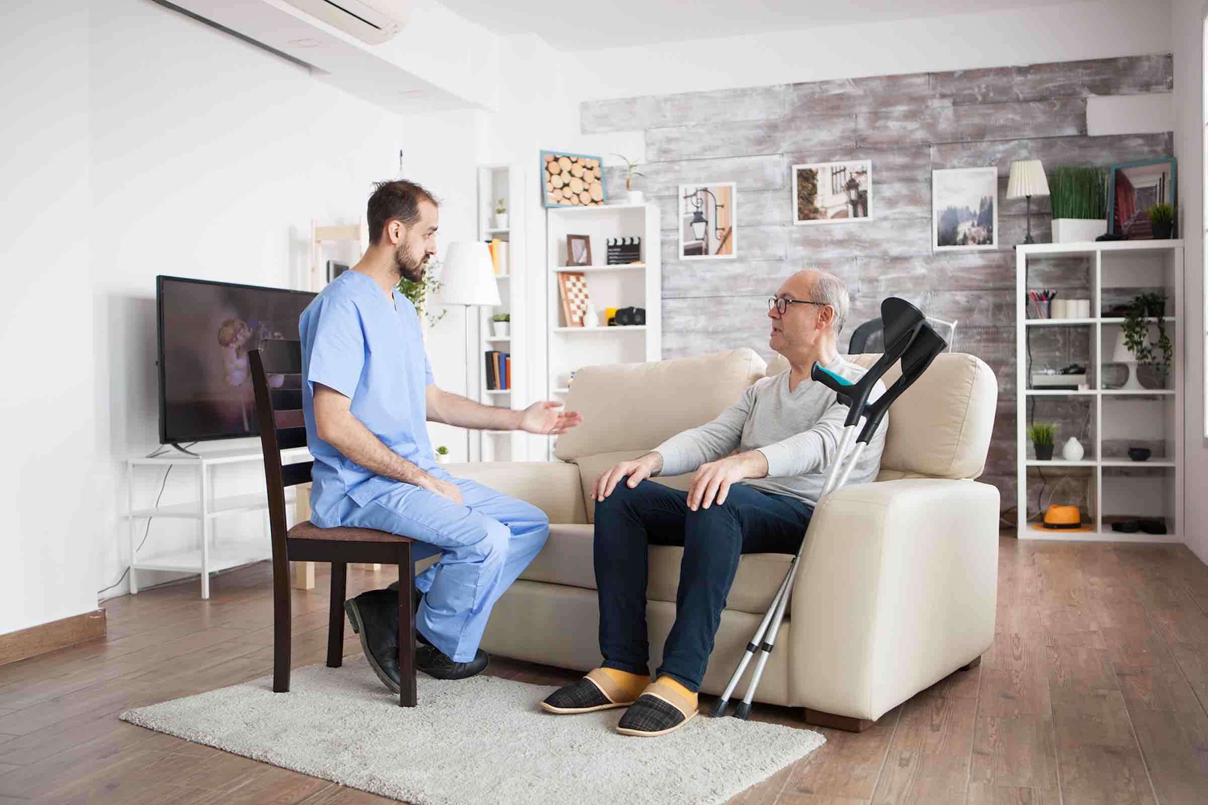 Male nurse on a chair in a nursing home talking with elderly age man while he is sitting on couch.